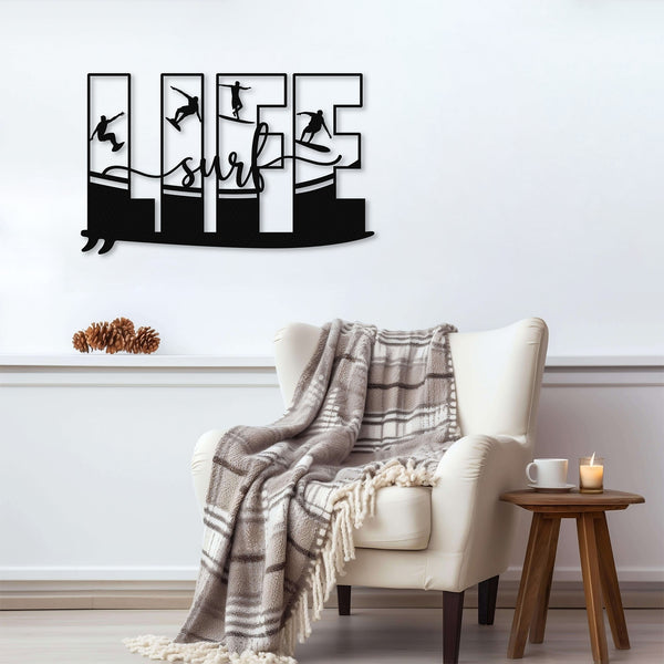 Black metal wall art featuring the word 'LIFE' with each letter containing a silhouette of a surfer in different poses. The word 'surf' is written in cursive below, with a stylized wave design connecting the letters riding on a surf board. The art is displayed on a light gray wall above a cozy armchair with a plaid blanket draped over it.