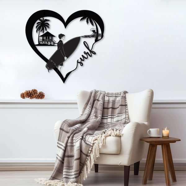 Black metal wall art featuring a surfer looking at riding a wave with a beach house, inside a heart shape with the word 'surf' written in cursive. The art is displayed on a white wall above a cozy armchair with a plaid blanket draped over it.