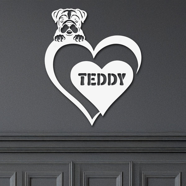 White metal sign with a dog silhouette peeking over a heart, with the name "Teddy" written inside the heart, hanging on a gray wall.