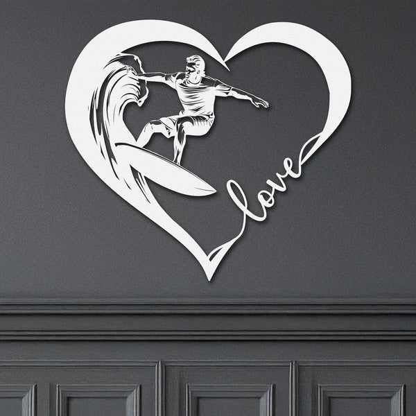 White metal wall art featuring a surfer riding a wave inside a heart shape with the word 'love' written in cursive. The art is displayed on a dark wall above a wainscotting.