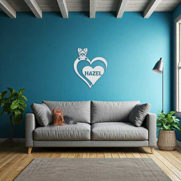 White metal sign with a Yorkshire Terrier silhouette peeking over a heart, with the name "Hazel" written inside the heart, hanging on a blue wall above a grey sofa with a Yorkie laying on it.