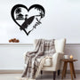 Black metal wall art featuring a surfer looking at riding a wave with a beach house, inside a heart shape with the word 'surf' written in cursive. The art is displayed on a white wall above a cozy armchair with a plaid blanket draped over it.