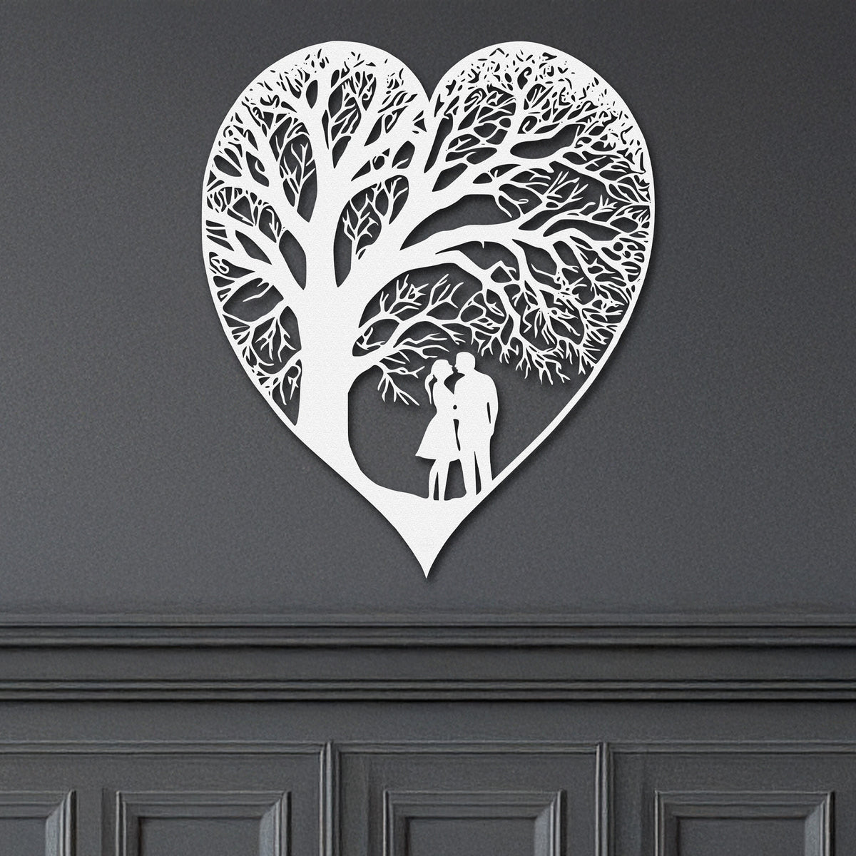 A white heart-shaped metal wall decor with a detailed tree-like pattern with a couple, representing the theme of everlasting love on a grey wall.