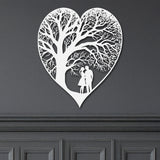 A white heart-shaped metal wall decor with a detailed tree-like pattern with a couple, representing the theme of everlasting love on a grey wall.