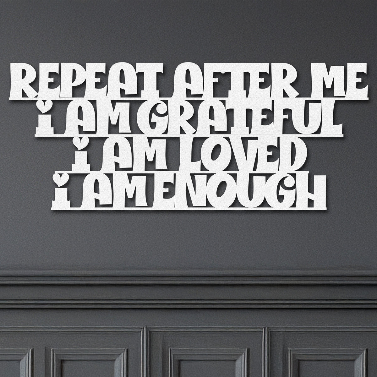 White metal die-cut sign with the phrase 'Repeat After Me I Am Grateful I Am Loved I Am Enough' mounted on a dark wall.