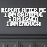 White metal die-cut sign with the phrase 'Repeat After Me I Am Grateful I Am Loved I Am Enough' mounted on a dark wall.