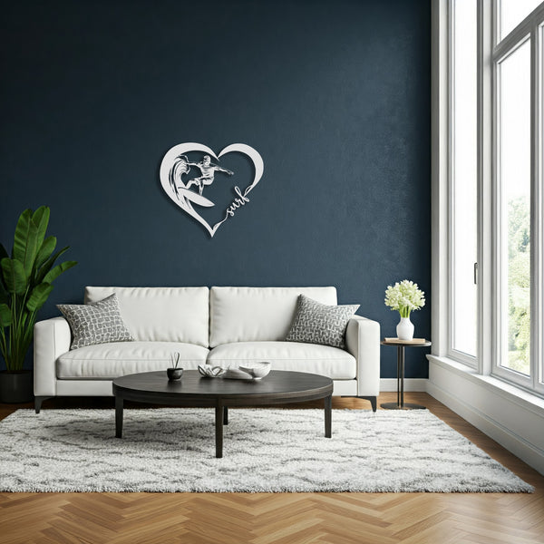 Silver metal wall art featuring a surfer riding a wave inside a heart shape with the word 'surf' written in cursive. The art is hung on a dark blue wall above a modern white sofa with decorative pillows.