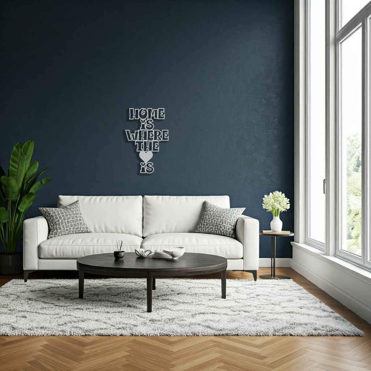 Silver metal sign with the phrase 'Home Is Where The Heart Is' displayed above a white sofa in a living room with large windows.