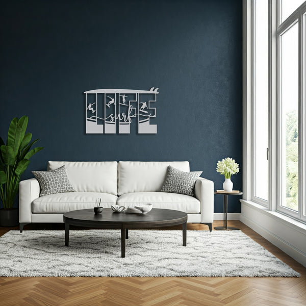 Decorative metal wall art featuring the word 'LIFE' with silhouettes of surfers in various poses within each letter, and the word 'surf' written in cursive script in the center hanging behind a white sofa in a living room with a large window.
