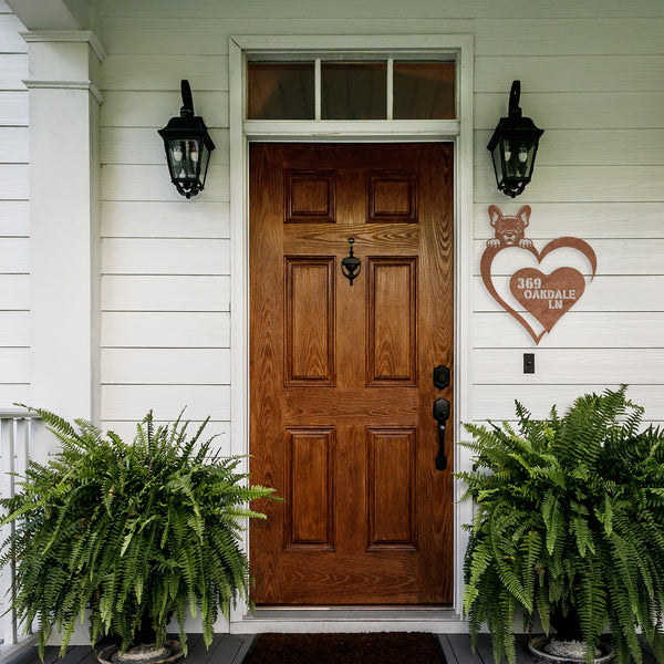 A wooden front door with a transom window above, flanked by two potted ferns. A custom metal sign featuring a heart shape with a dog peeking over the top and the address '369 Oakdale LN' is mounted to the right of the door.