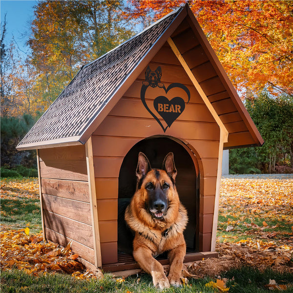 A German Shepherd dog sitting in a wooden doghouse with a custom metal sign above the entrance featuring a heart shape and the name 'BEAR'