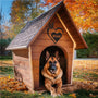 A German Shepherd dog sitting in a wooden doghouse with a custom metal sign above the entrance featuring a heart shape and the name 'BEAR'