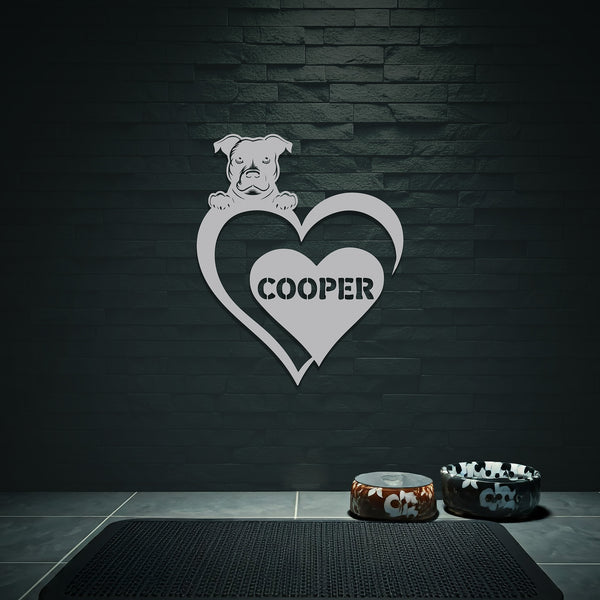 Silver metal sign with a dog silhouette peeking over a heart, with the name "C ooper" written inside the heart, hanging on a grey wall above dog bowls.
