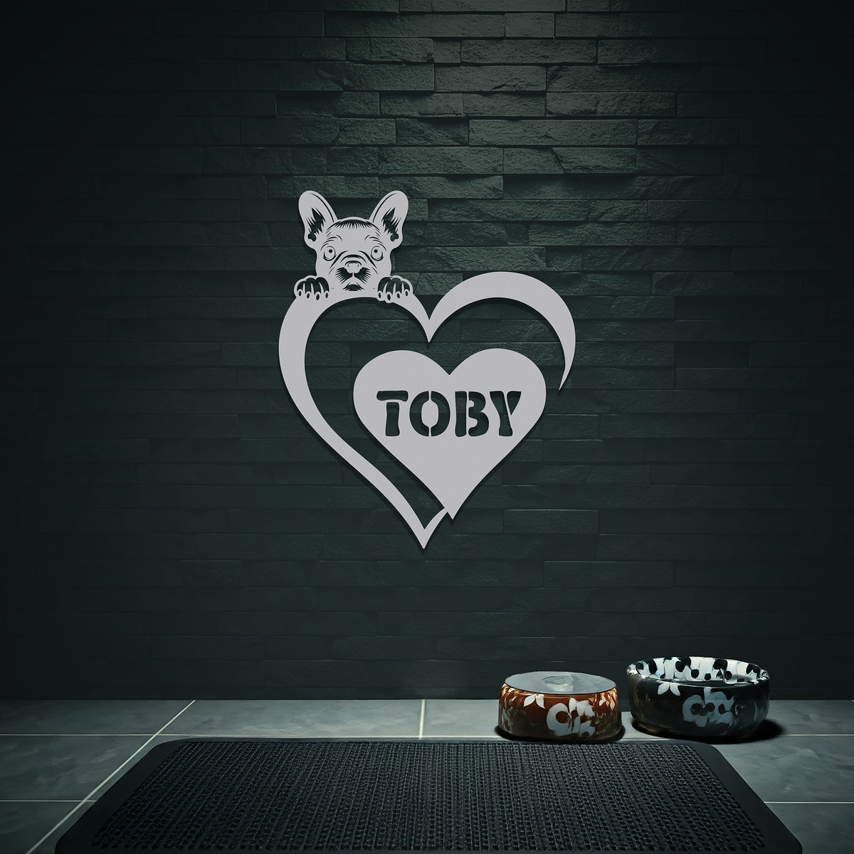 Silver metal sign with a dog silhouette peeking over a heart, with the name "Toby" written inside the heart, hanging on a grey wall above dog bowls.