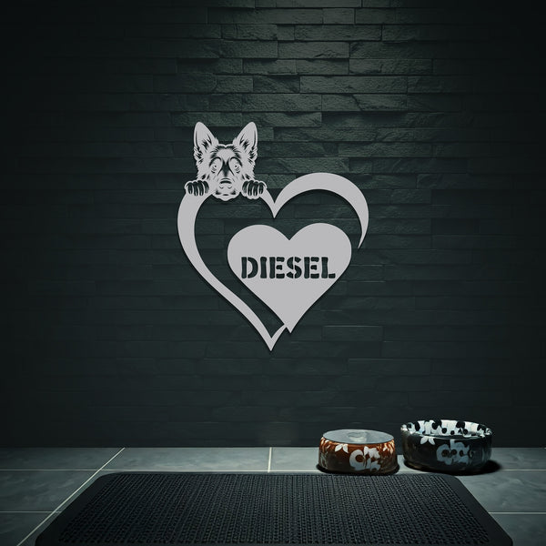 Silver metal sign of a German Shepard dog silhouette peeking over a heart, with the name "Diesel" written inside the heart, hanging on a gray wall.