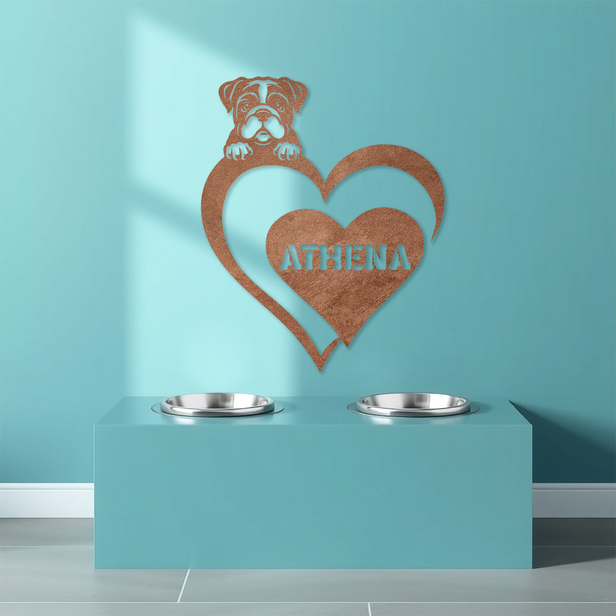 Copper colored metal sign with a dog silhouette peeking over a heart, with the name "Athena" written inside the heart, hanging on a blue wall above dog bowls.